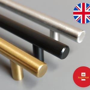 May include: Close-up of three cabinet pulls in gold, black, and silver. The cylindrical pulls are arranged diagonally, showcasing their different finishes. The gold pull has a brushed texture, while the black and silver pulls have a smooth finish. The pulls are for cabinets and drawers.