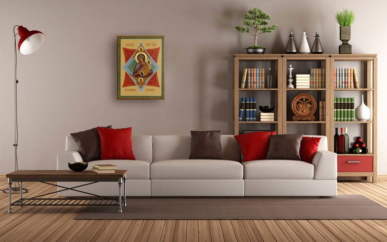 K&ouml;nnte beinhalten: Ein wei&szlig;es Sofa mit roten und braunen Kissen steht in einem Wohnzimmer mit einem B&uuml;cherregal aus Holz, das mit B&uuml;chern und Dekorationsgegenst&auml;nden gef&uuml;llt ist. &Uuml;ber dem Sofa h&auml;ngt ein gerahmtes religi&ouml;ses Symbol.