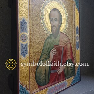 May include: A hand-painted icon of Saint Paul, depicted with a book and a halo. The icon is framed in a gold-leaf border with intricate floral designs. The background is a rich gold colour.