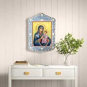 May include: A framed religious icon depicting the Virgin Mary holding a baby Jesus. The frame is white with blue and white floral designs. The icon is set against a white wood panel background.