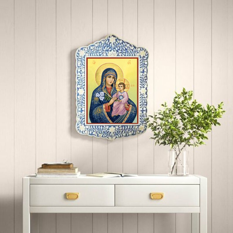 May include: A framed religious icon depicting the Virgin Mary holding a baby Jesus. The frame is white with blue and white floral designs. The icon is set against a white wood panel background.