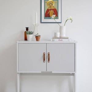 May include: A white cabinet with two doors and brown leather handles. The cabinet is on a white floor and has a white wall behind it. There is a framed picture of a religious icon above the cabinet, and a vase with white flowers on the top of the cabinet.