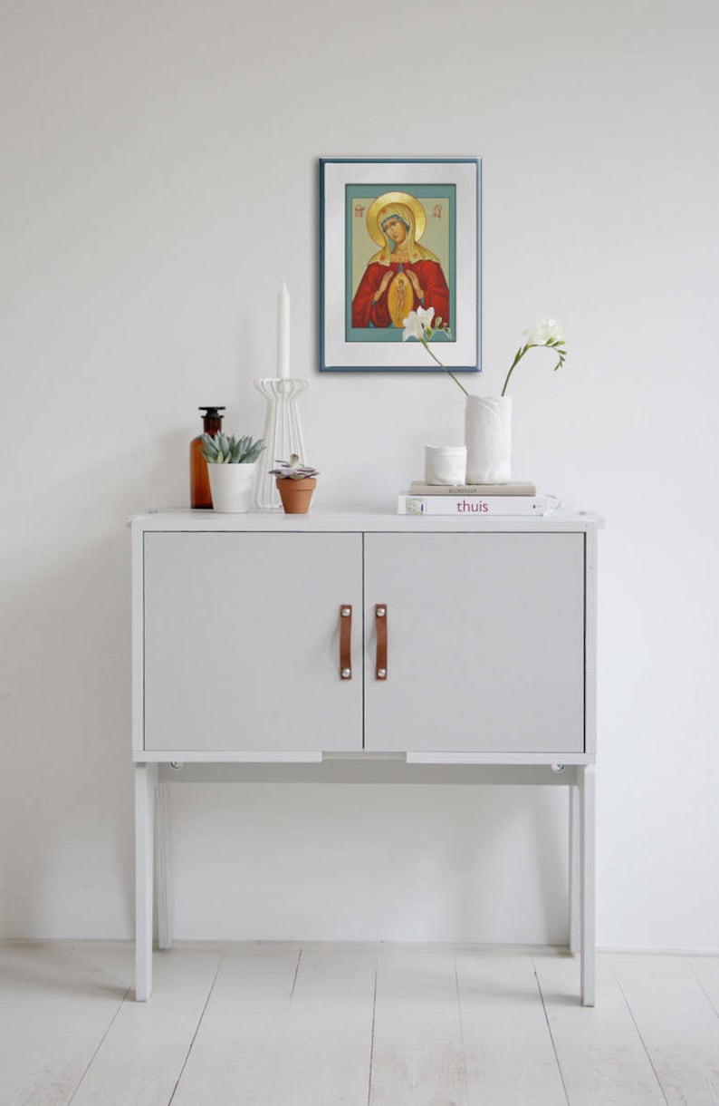 May include: A white cabinet with two doors and brown leather handles. The cabinet is on a white floor and has a white wall behind it. There is a framed picture of a religious icon above the cabinet, and a vase with white flowers on the top of the cabinet.