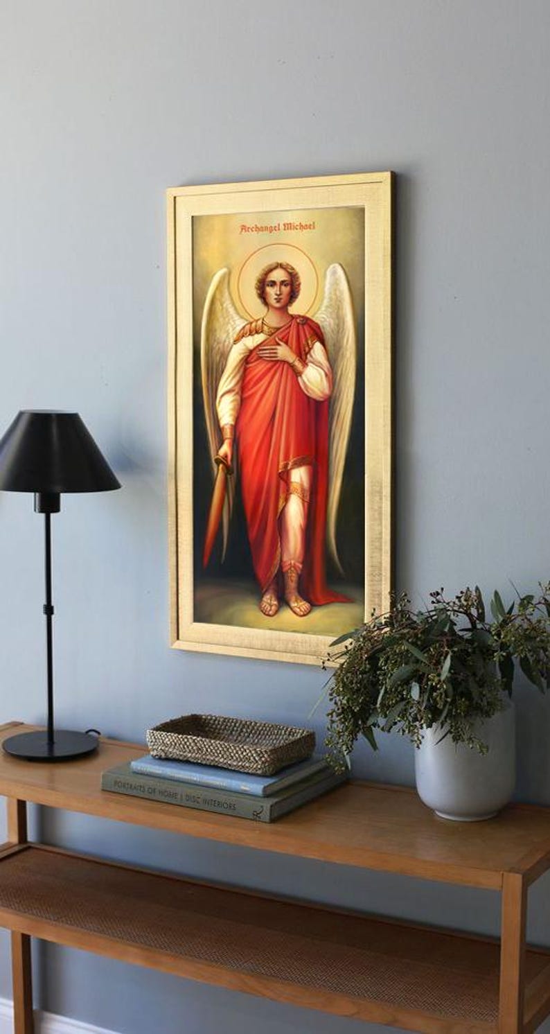 Pode incluir: Uma pintura emoldurada do Arcanjo Miguel, um &iacute;cone religioso com uma aur&eacute;ola dourada, segurando uma espada e vestindo uma t&uacute;nica vermelha e uma armadura branca. A pintura est&aacute; em uma prateleira de madeira com livros e uma cesta de vime.