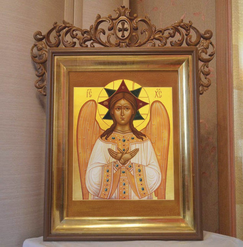 May include: A framed icon depicting an angel with wings, wearing a white robe and a gold crown. The angel has a halo with a star and the letters "IC XC" in the background. The frame is ornate and has a gold finish.