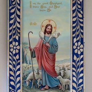 May include: A framed painting of Jesus Christ as the Good Shepherd. He is depicted with a lamb on his shoulders and a staff in his hand. The painting is set against a blue sky with white clouds and a green field with sheep. The frame is made of white bone with a blue background and a floral design.