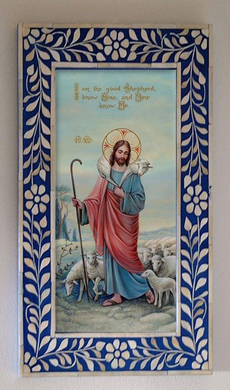 May include: A framed painting of Jesus Christ as the Good Shepherd. He is depicted with a lamb on his shoulders and a staff in his hand. The painting is set against a blue sky with white clouds and a green field with sheep. The frame is made of white bone with a blue background and a floral design.