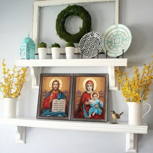 May include: A white shelf with two framed religious paintings, a teal lantern, three white ceramic pots with green plants, two decorative plates, and two vases with yellow flowers. The shelf is mounted on a white wall.