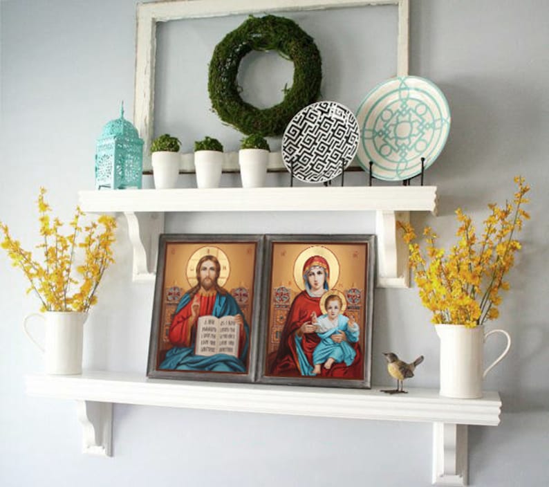 May include: A white shelf with two framed religious paintings, a teal lantern, three white ceramic pots with green plants, two decorative plates, and two vases with yellow flowers. The shelf is mounted on a white wall.