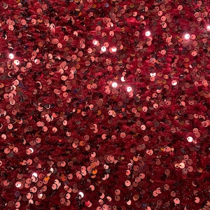 May include: A close-up shot of a deep red fabric covered in small, round sequins. The sequins are scattered across the surface, creating a shimmering effect. The fabric has a soft, velvety texture.