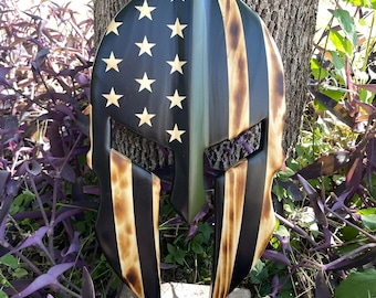 May include: Wooden Spartan helmet with an American flag design. The helmet has a black and tan finish with gold stars and vertical stripes. It features a green stripe. The helmet is hanging on a tree.