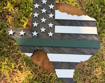 May include: A wooden cutout shaped like the state of Texas, featuring a dark grey stained finish with white horizontal stripes and a single horizontal green stripe. The design includes several small, raised white stars on the left side.