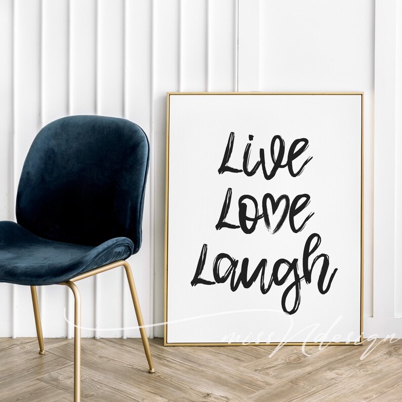 May include: A framed print with the words "Live Love Laugh" in black brush lettering. A navy blue velvet chair with gold legs is to the left of the print. The background is a white wall and wooden floor.
