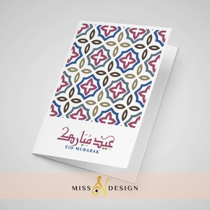 May include: A white card with a geometric pattern in shades of blue, brown, and pink. The card has the text "Eid Mubarak" in Arabic script and the English translation below. The card is folded in half and has a white background.