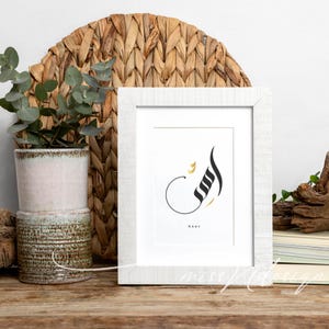 Puede incluir: Impresi&oacute;n art&iacute;stica enmarcada en blanco con caligraf&iacute;a &aacute;rabe en negro y dorado. La impresi&oacute;n se muestra sobre una superficie de madera, con un tapiz de pared tejido y una planta en maceta en el fondo. La palabra "Aser" est&aacute; escrita debajo.