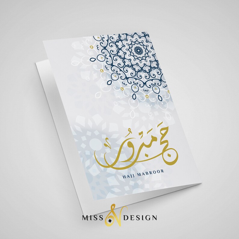 May include: A white card with a blue and gold design. The card has a mandala pattern in the top right corner and Arabic calligraphy in gold in the center. The text "Hajj Mabroor" is written in English below the calligraphy.