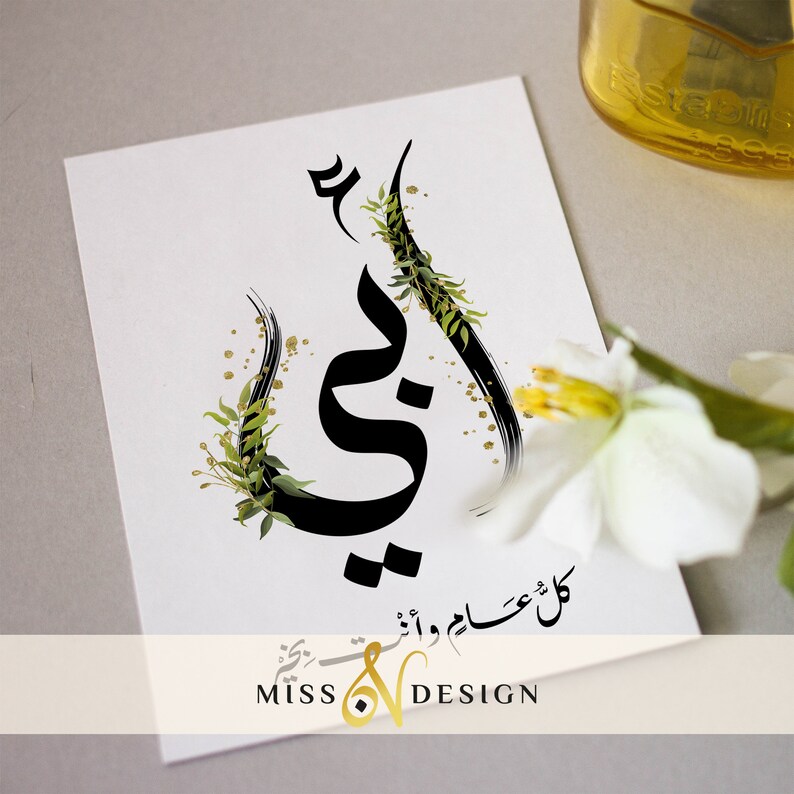 May include: A white card with black calligraphy that says "Mama" in Arabic script. The calligraphy is surrounded by green leaves and gold glitter. The card also has the text "كل عام وانت بخير" in Arabic script, which translates to "Happy New Year". The card is signed "Miss & Design".
