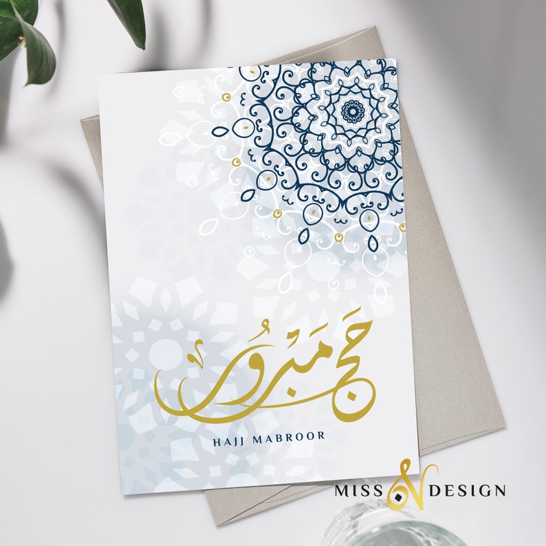 May include: A white card with a blue and white mandala design in the top right corner. The card has gold Arabic calligraphy that says "Hajj Mabroor" in the center. The card is on a gray surface.