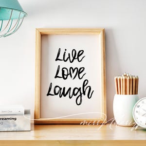 May include: A framed print with the words "Live Love Laugh" in black script on a white background. The frame is made of wood. A white vase with pencils and a clock are on the wooden surface.