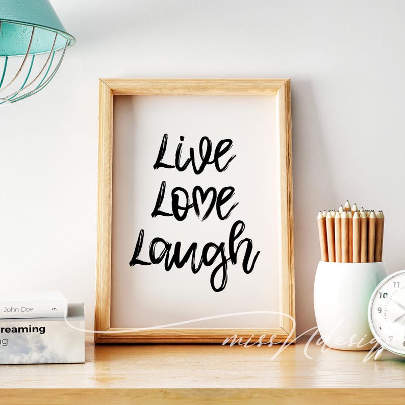 May include: A framed print with the words "Live Love Laugh" in black script on a white background. The frame is made of wood. A white vase with pencils and a clock are on the wooden surface.