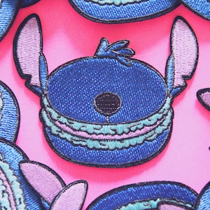 Peut inclure: Patch brodé bleu de Stitch de Lilo & Stitch, portant un macaron bleu et rose. Le patch a un fond rose.