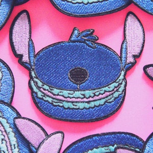 May include: A collection of embroidered patches featuring a blue character shaped like a macaron. The design includes dark blue rounded edges, a light blue and pink ruffled center, a black circular nose, and two tall, pointed purple-lined ears.