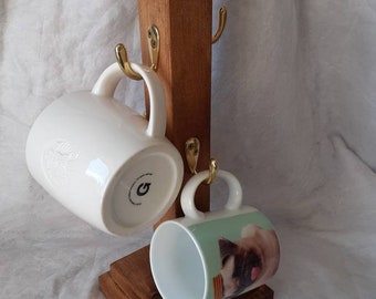 May include: A wooden mug rack with four gold hooks. Two mugs are hanging on the hooks. One mug is white with a design on it. The other mug is white with a green background and a picture of a pug on it.