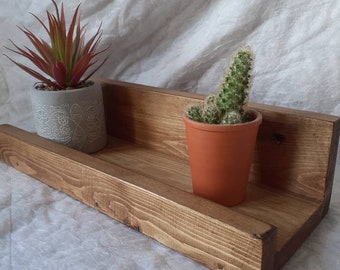 Puede incluir: Una estantería de madera con dos plantas en maceta. La estantería está teñida de un color marrón oscuro y tiene un aspecto rústico. Las plantas son una suculenta y un cactus.