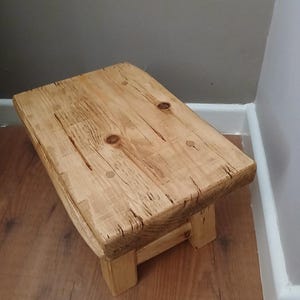Może przedstawiać: Mały, rustykalny drewniany stołek z prostokątnym siedziskiem i czterema solidnymi nogami. Drewno ma naturalny, jasnobrązowy kolor z widocznym usłojeniem i sękami, co nadaje mu ręcznie robiony wygląd. Stołek ma około 30 cm długości, 20 cm szerokości i 25 cm wysokości.