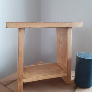 Puede incluir: Una pequeña mesa auxiliar de madera marrón claro con un estante inferior. La mesa tiene cuatro patas y una parte superior rectangular. La madera tiene un acabado natural. La mesa mide aproximadamente 61 cm de altura.