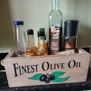 Może przedstawiać: Drewniana skrzynka z napisem "Finest Olive Oil" i grafiką oliwek. Zawiera butelki z olejem, octem i przyprawami, w tym butelkę octu balsamicznego i młynek do pieprzu.