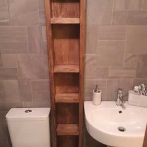 May include: A tall, narrow wooden bathroom shelf unit with four shelves. The unit is made of light brown wood and is positioned next to a white pedestal sink and a toilet. The shelves are open and designed for storage.