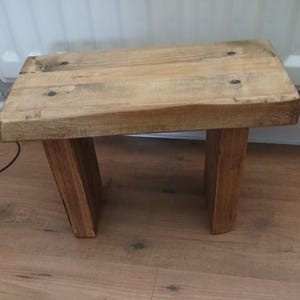 May include: A small, rustic wooden stool. It has a rectangular top and four sturdy legs. The stool is made of light-coloured wood with visible grain and knots, giving it a natural, handcrafted appearance. The legs are angled for stability.