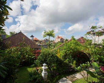 May include: A view of a lush green garden with a tiled roof building in the background. The garden has a variety of plants and trees, including a palm tree. The sky is cloudy and blue.