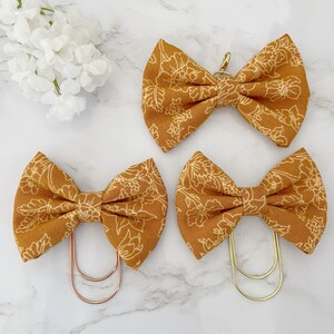 May include: Three mustard yellow fabric bow clips with a white floral pattern. The bow clips are made of fabric and have a metal clip on the back.