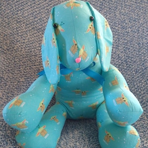 May include: A soft toy bunny made from light blue fabric, decorated with a repeating cartoon character pattern. The bunny features long ears, black button eyes, and a pink nose. A blue ribbon is tied around its neck.