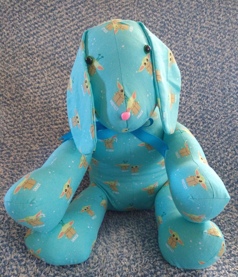May include: A soft toy bunny made from light blue fabric, decorated with a repeating cartoon character pattern. The bunny features long ears, black button eyes, and a pink nose. A blue ribbon is tied around its neck.