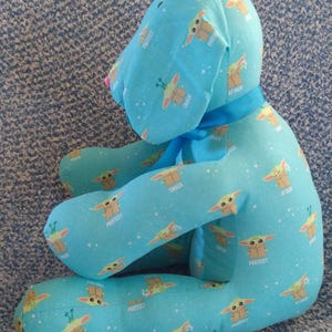 May include: A light blue plush dog-shaped pillow with a repeating pattern of a cartoon character and the words "Protect", "Snack", and "Attack". It has a blue ribbon and a pink nose. The pillow is designed to be a decorative item.