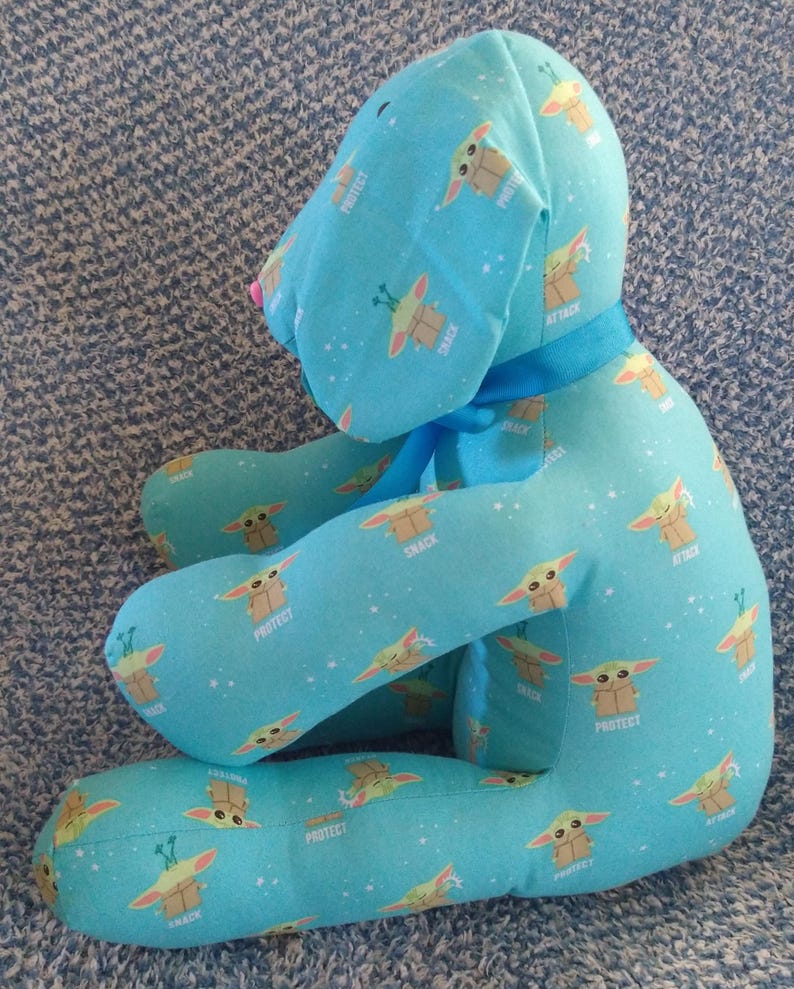 May include: A light blue plush dog-shaped pillow with a repeating pattern of a cartoon character and the words "Protect", "Snack", and "Attack". It has a blue ribbon and a pink nose. The pillow is designed to be a decorative item.