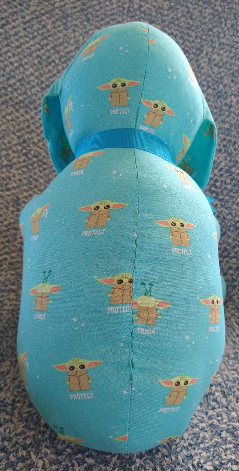 May include: A light blue plush toy featuring a repeating pattern of a character, the word "PROTECT", and the word "SNACK". The toy has a blue ribbon around its neck. The toy is a character from a popular science fiction series.