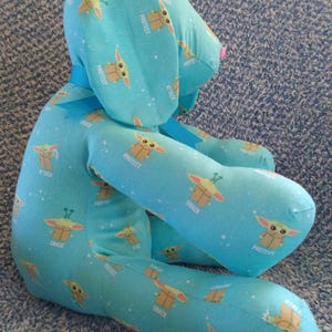 May include: A soft toy dog in a seated pose, made from light blue fabric printed with a cartoon character and the words "PROTECT", "ATTACK", and "SNACK". The dog has a pink nose, green eyes, and a blue ribbon.
