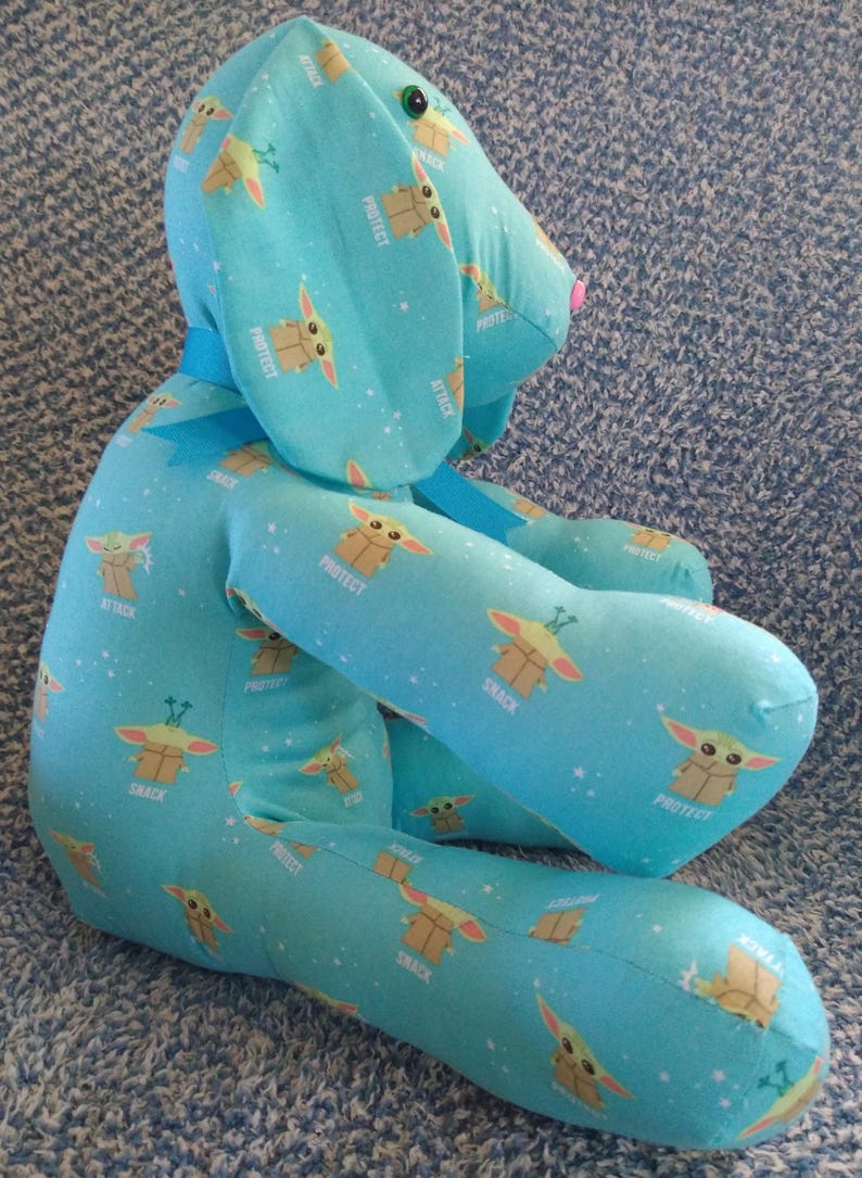 May include: A soft toy dog in a seated pose, made from light blue fabric printed with a cartoon character and the words "PROTECT", "ATTACK", and "SNACK". The dog has a pink nose, green eyes, and a blue ribbon.