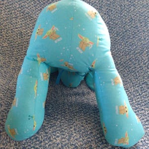 May include: A light blue reading pillow with a cartoon character print. The pillow has a repeating pattern of a cartoon character with the words "ATTACK" and "PROTECT" in white. The pillow has two leg-like extensions.