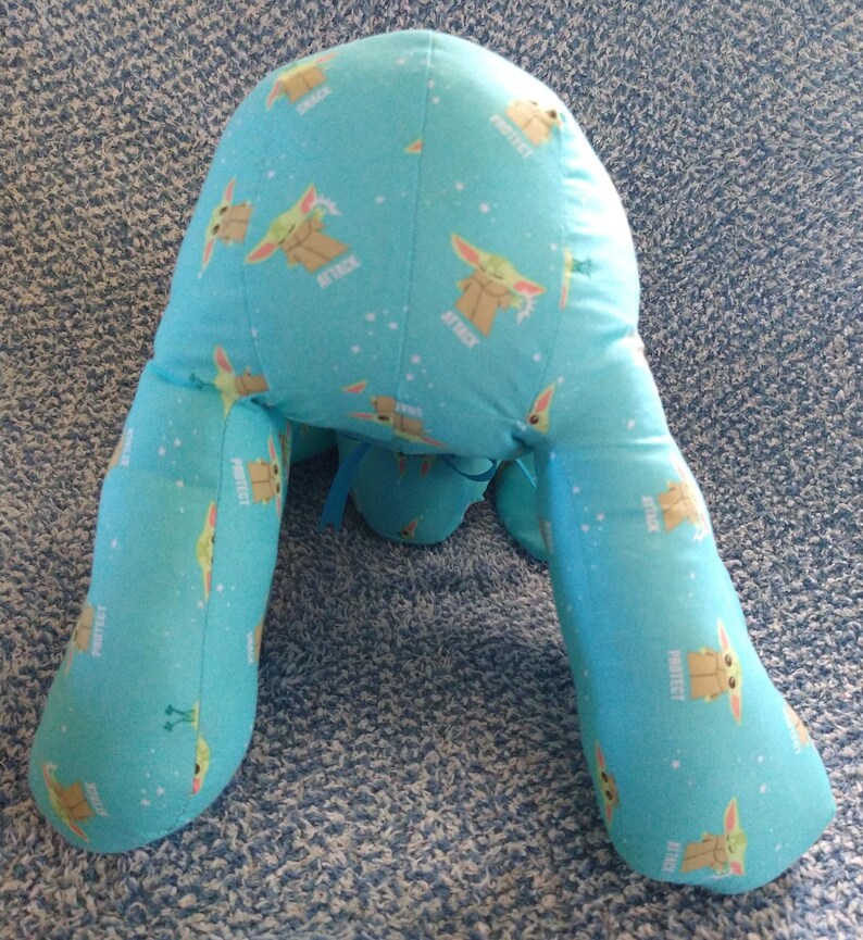 May include: A light blue reading pillow with a cartoon character print. The pillow has a repeating pattern of a cartoon character with the words "ATTACK" and "PROTECT" in white. The pillow has two leg-like extensions.