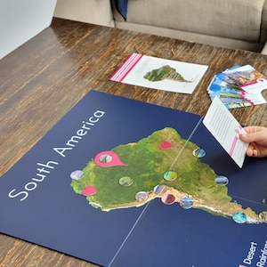 May include: A child is playing with a South America map game on a wooden table. The map is blue with a green illustration of the continent and pink location markers. There are cards and circular game pieces scattered around.