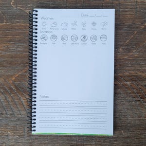 May include: A spiral-bound notebook with a white cover featuring weather and location icons. The notebook includes sections for the date, weather conditions, location, and notes. The notebook is open on a wooden surface.