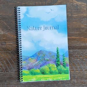 May include: A spiral-bound "Nature Journal" notebook. The cover features a watercolor painting of a landscape with mountains, trees, and a blue sky with clouds. The journal is on a wooden surface.