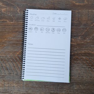 May include: A spiral-bound notebook with a white cover and lined pages. The top of the page has sections for weather, date, and location, with corresponding icons. The notebook is open, revealing the interior pages and a green edge.