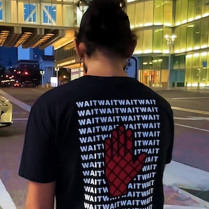 以下が含まれることがあります： 赤い手のグラフィックと「WAITWAITWAITWAIT」というテキストが複数回繰り返された黒いTシャツです。