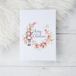 May include: A white Christmas card with a watercolour illustration. It features a pink and gold nutcracker soldier, surrounded by a floral wreath with the words "Merry Christmas". Small wrapped gifts are at the bottom of the card.